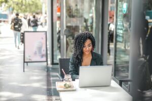 High angle of pensive African American female freelancer in glasses and casual clothes focusing on screen and interacting with netbook while sitting at table with glass of yummy drink on cafe terrace in sunny day